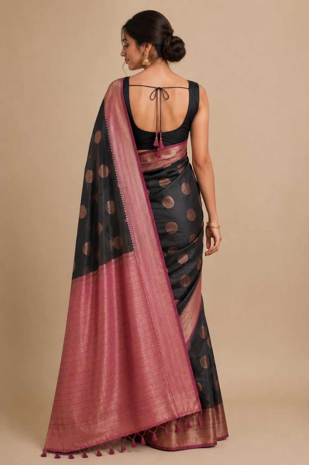 A woman shown from the back wearing a black and dusty pink saree with gold motifs, paired with a black sleeveless blouse, against a solid beige studio background. 