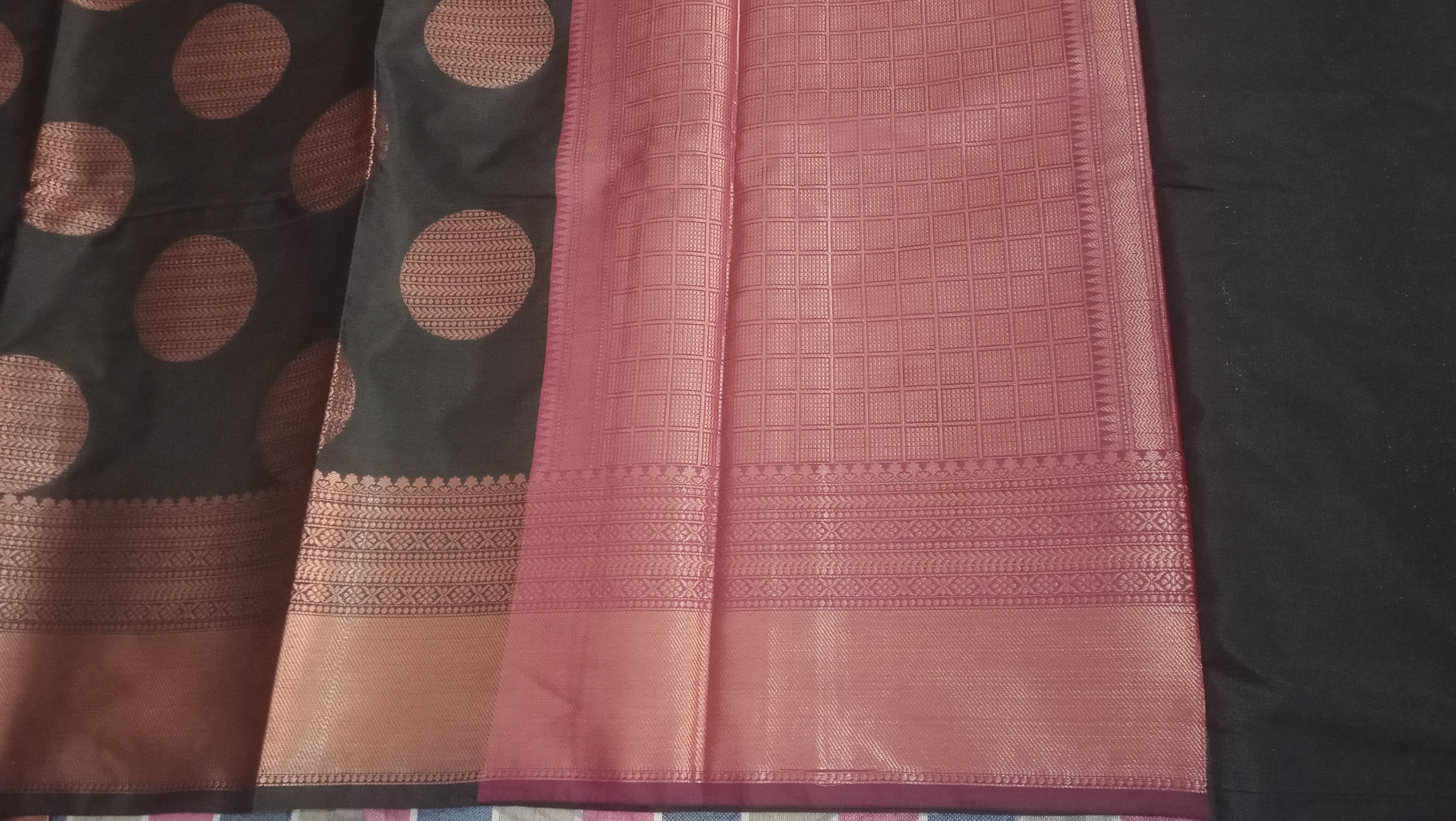 A close-up of a folded textile  saree, showing a black body with large metallic bronze-gold circular motifs and a wide pink border with woven geometric detailing.