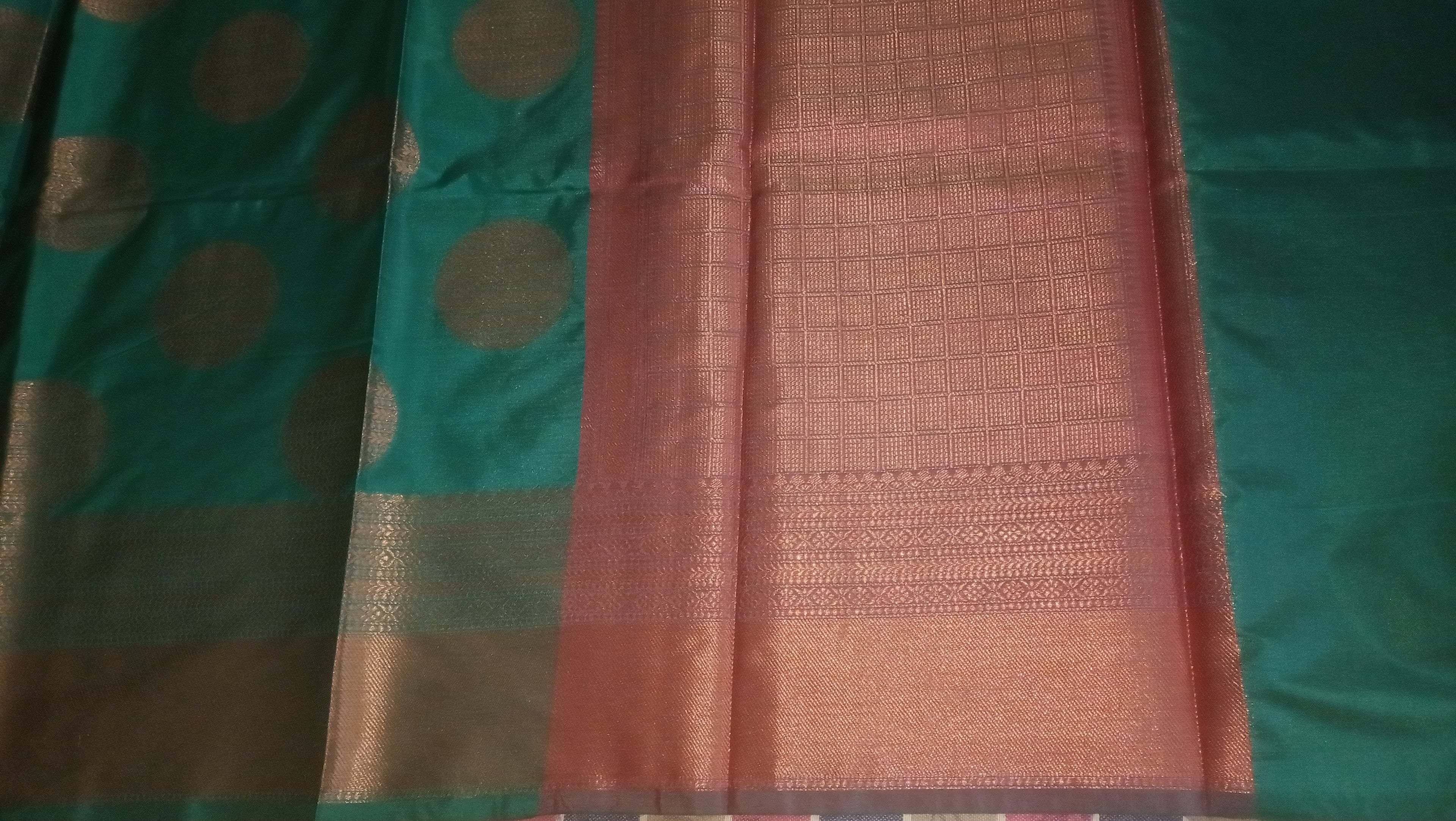 A close-up of a woven textile a sari, featuring a green body with metallic gold circular motifs, central panel with gold woven detailing, and a teal green border. 