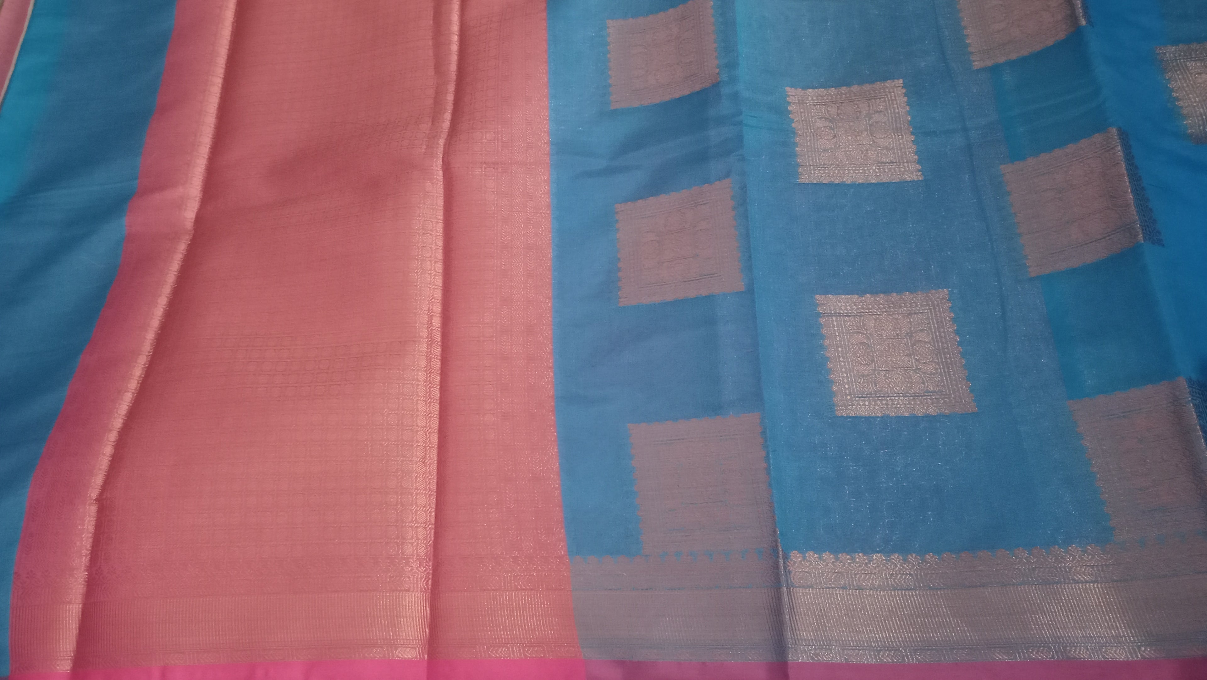 A close-up of a textile fabric with a blue body, a turquoise pink border, and woven metallic silver geometric patterns.