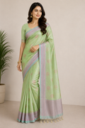A woman modeling a light green saree with lavender and blue borders, featuring small gold motifs and a matching blouse, set against a neutral beige background with a potted plant on the left.
