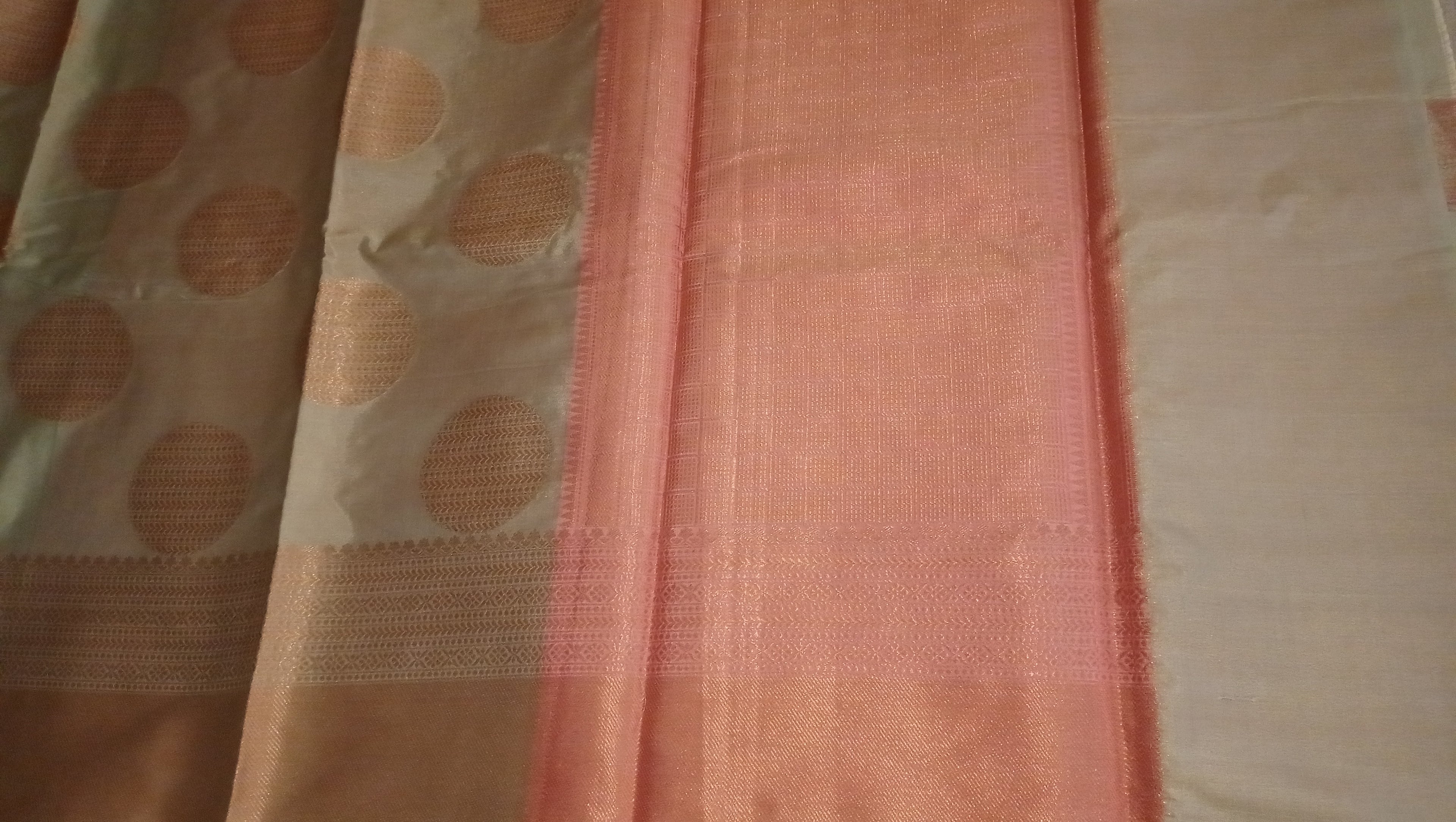 A close-up of a draped fabric, likely a saree, showing a peach-pink border, a beige body with gold circular motifs, and a silver shining border. 