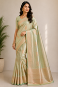 A woman modeling a light sage green silk saree with a shimmering gold border and pallu, set against a neutral beige background with a potted plant on the left.