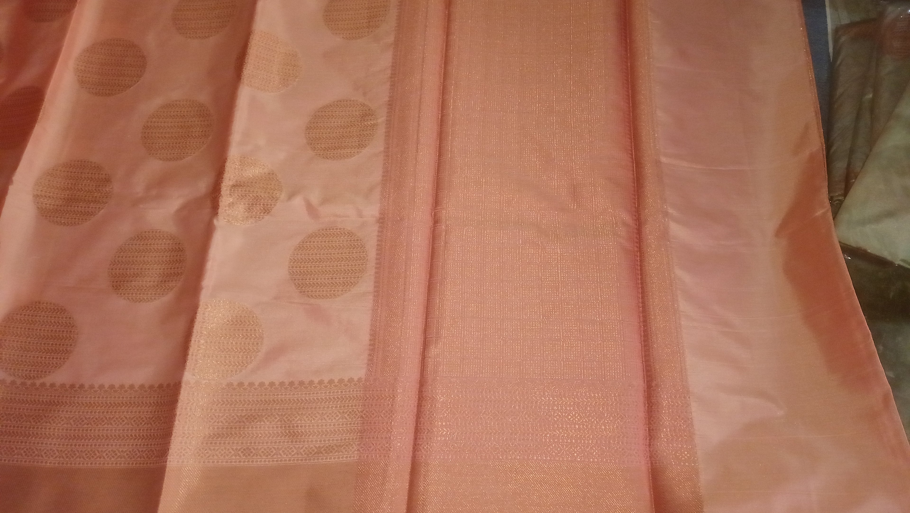A peach-pink silk saree with a wide border featuring large gold circular motifs, shown folded. 