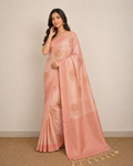 A woman modeling a peach-pink saree with gold floral motifs and a matching border, standing against a neutral beige wall and textured floor.