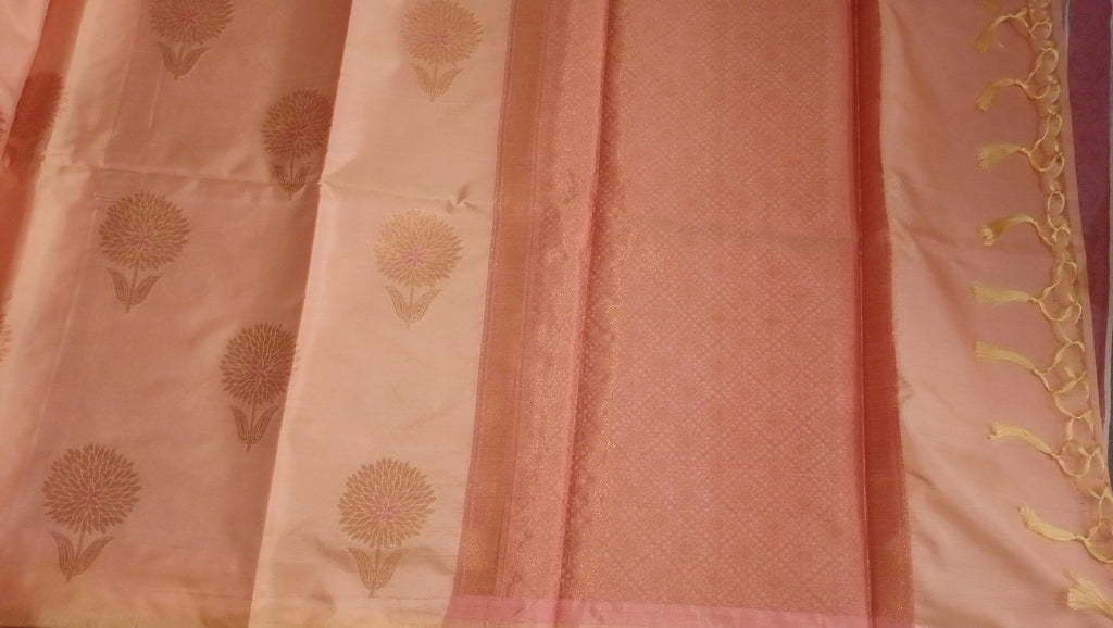 A close-up of a pink and peach fabric textile, likely a sari, featuring a solid pink border, a lighter peach body with gold floral motifs, and a decorative gold tassel along the edge.