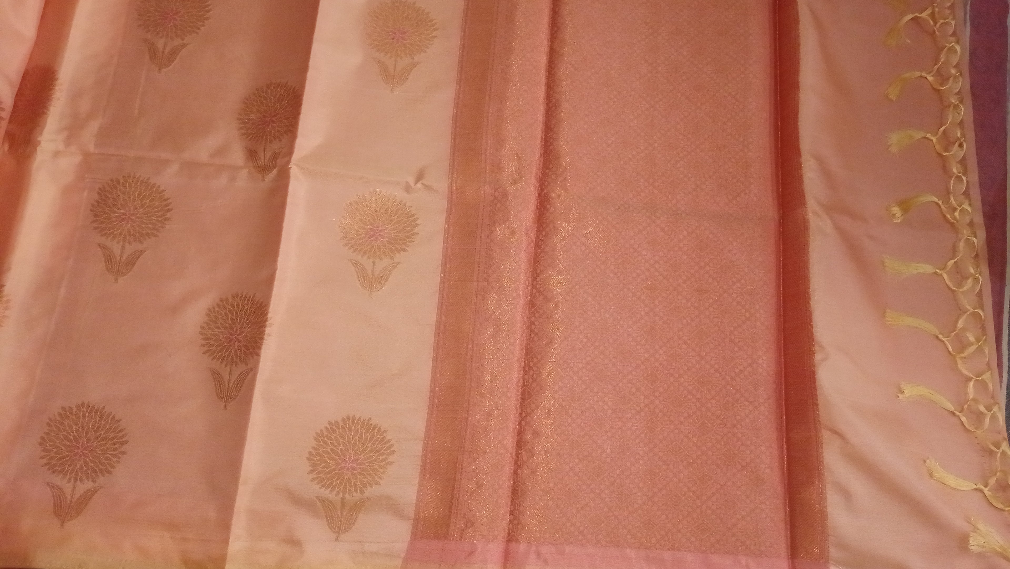A close-up of a pink and peach fabric textile, likely a sari, featuring a solid pink border, a lighter peach body with gold floral motifs, and a decorative gold tassel along the edge.