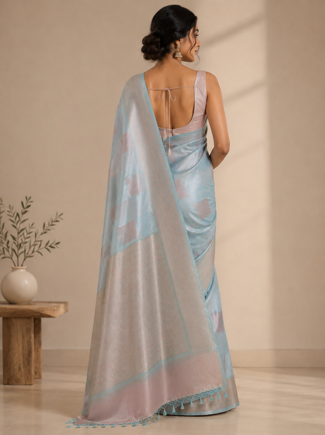 A woman wearing a light blue saree, shown from the back in a warm beige setting.